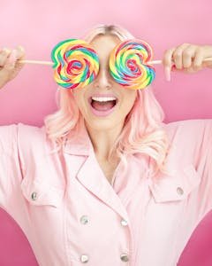 Creative portrait of a joyful woman with pink hair holding vibrant lollipops against a pastel pink background.