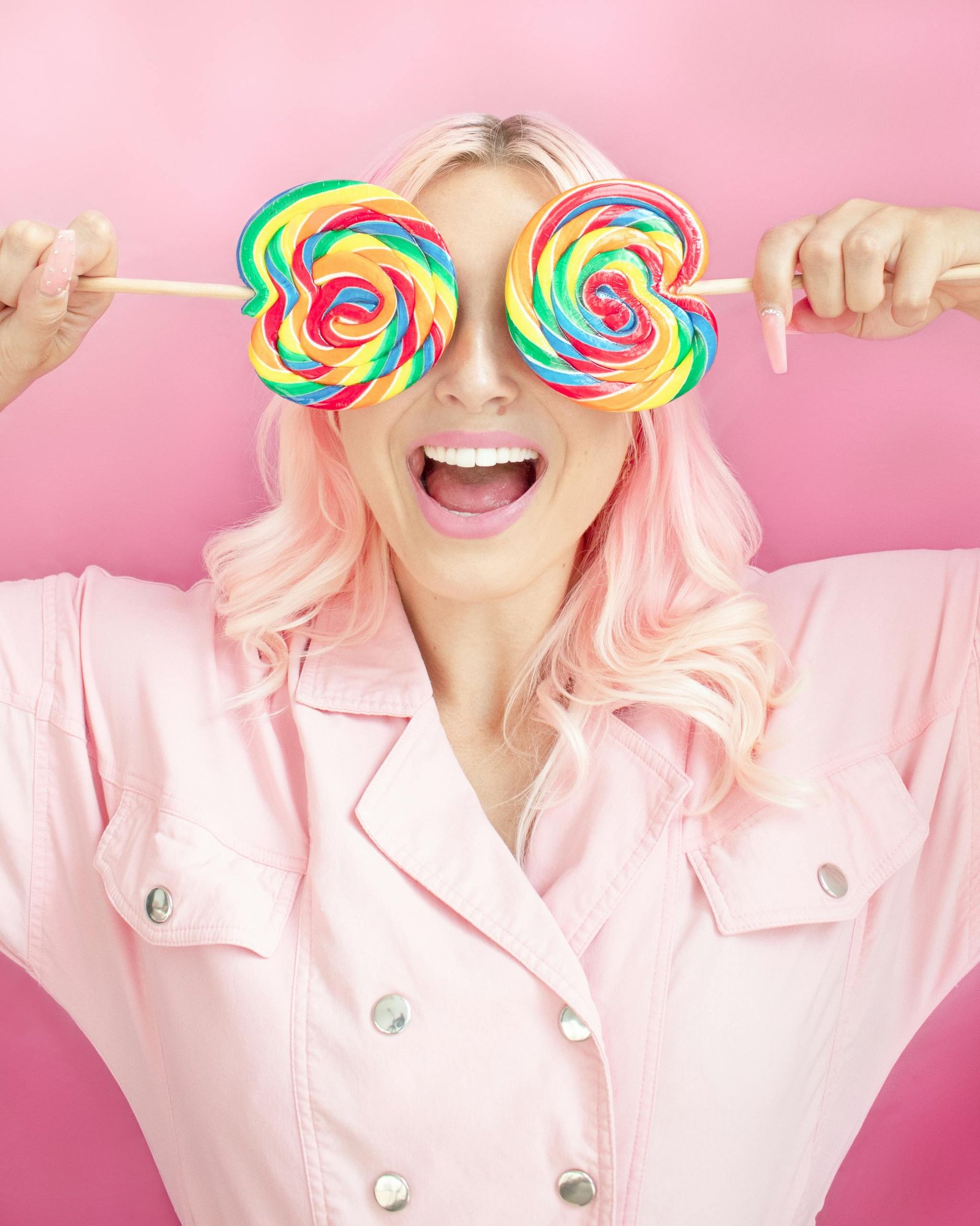 Creative portrait of a joyful woman with pink hair holding vibrant lollipops against a pastel pink background.