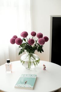 Beautiful pink flowers in a glass vase, accompanied by a book and perfume on a white tabletop.